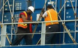 Two people with orange vest on a crane next to blue containers